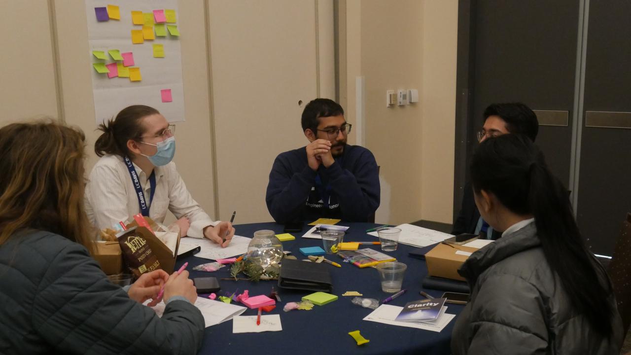 A group of five people sitting around a table engaged in discussion during a workshop session. The table is covered with a blue tablecloth and scattered with sticky notes, pens, paper and snacks. One participant is wearing a face mask and taking notes, while others listen and converse. A large sheet of paper with colorful sticky notes is attached to the wall in the background.